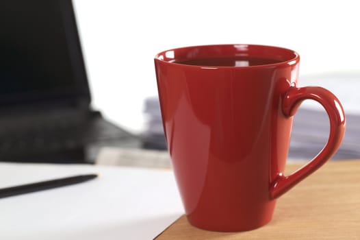 Red coffee cup standing on desk with blank paper and pen, with laptop and staple of papers in back (Selective Focus, Focus on the rim of the cup) 