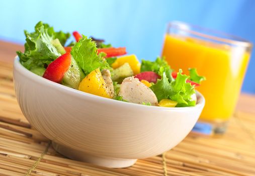 Fresh and healthy chicken salad with lettuce, mango, red bell pepper and cucumber with fresh juice in the back in front of blue background (Selective Focus, Focus on the front of the salad)