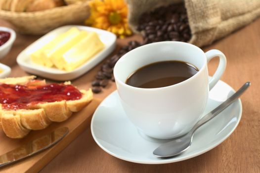 Hot, fresh coffee with croissant with strawberry jam, butter and coffee beans in the back (Selective Focus, Focus on the front rim of the coffee cup)