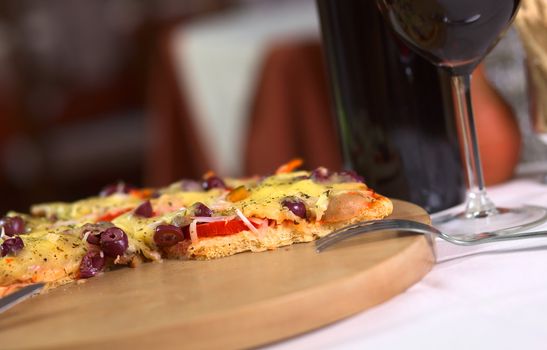 A partly eaten pizza on wooden plate on a table with wine in a restaurant (Selective Focus, Focus on the front of the pizza)