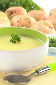 Fresh potato cream soup garnished with a parsley leaf with green onion, baguette slices, raw potatoes and parsley in the back (Selective Focus, Focus on the parsley leaf in the bowl) 