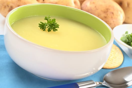 Fresh potato cream soup garnished with a parsley leaf with green onion and raw potatoes in the back on a blue table mat (Selective Focus, Focus on the parsley leaf in the bowl) 