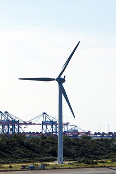 Large modern windmill with industrial cranes in background and small cars and people at the feet