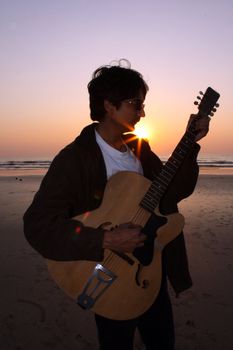 A young Indian guitarist performing on a beach at sunset.