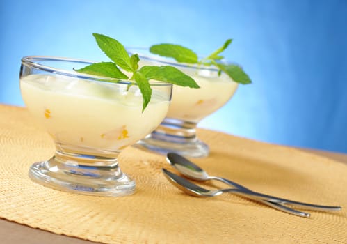 Mango cream cheese dessert with mint in glass bowl with a dessert spoons on yellow table mat with blue background (Selective Focus, Focus on the front of the glass and the leaves)