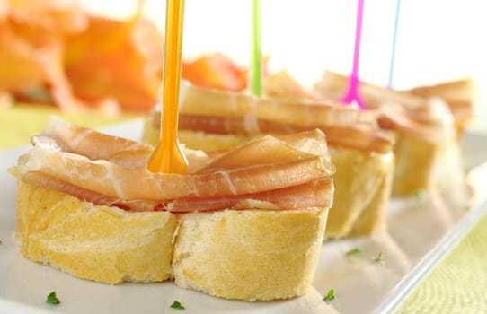 Baguette pieces with thin ham slices on top on plastic skewer served on a long white plate with a gladiolus flower in the back (Selective Focus, Focus on front)