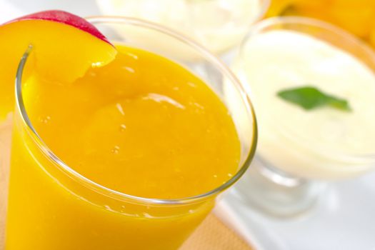 Fresh mango smoothie with cream cheese desserts in the background (Selective Focus, Focus on the front of the glass rim)
