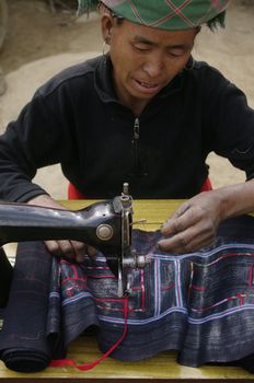Black Hmong woman at her sewing machine. It is installed in front of his home. His hands are blue dyeing fabrics she sews. This machine is the sole property of the couple.