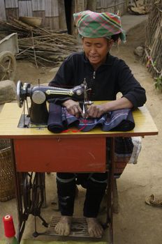 Black Hmong woman at her sewing machine. It is installed in front of his home. His hands are blue dyeing fabrics she sews. This machine is the sole property of the couple