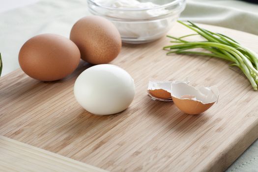 Peeled hard boiled egg with shells, two whole eggs and chives on cutting board.