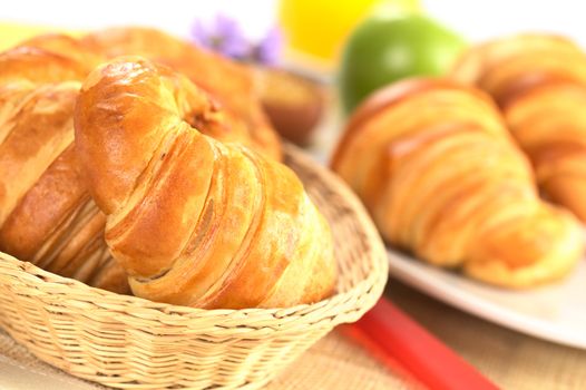 Fresh croissants in bread basket with a red knife beside, and a plate with croissants, green apple and orange juice in the back (Selective Focus, Focus on the front of the croissant in the basket)