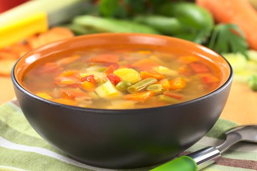 Fresh vegetable soup made of green bean, pea, carrot, potato, red bell pepper, tomato and leek in black bowl with ingredients in the back (Selective Focus, Focus on the vegetables in the middle of the soup)