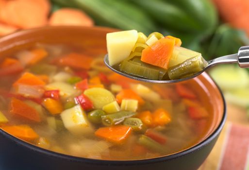 Fresh vegetable soup on spoon made of green bean, pea, carrot, potato, red bell pepper, tomato and leek in black bowl with ingredients in the back (Selective Focus, Focus on the carrot, leek and green bean on the spoon)