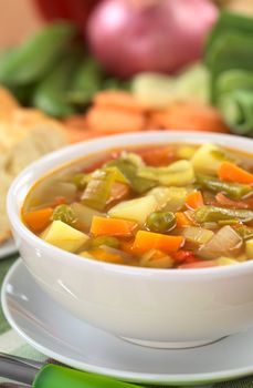 Fresh vegetable soup made of green bean, pea, carrot, potato, red bell pepper, tomato and leek in white bowl with baguette slices and ingredients in the back (Selective Focus, Focus on the vegetables one third into the soup)