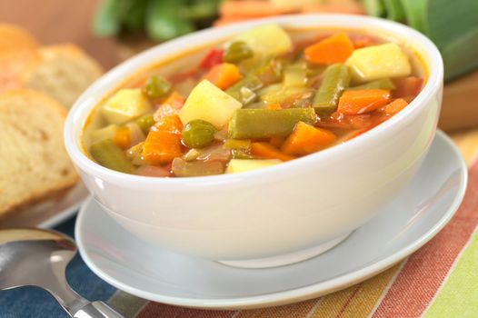 Fresh vegetable soup made of green bean, pea, carrot, potato, red bell pepper, tomato and leek in white bowl with baguette slices and ingredients in the back (Selective Focus, Focus on the vegetables one third into the soup)