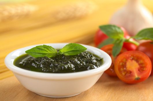 Fresh pesto made of basil, garlic and olive oil in a small bowl and garnished with a basil leaf with raw spaghetti, cherry tomatoes, basil leaves and garlic in the back (Selective Focus, Focus on the basil leaf in the bowl)