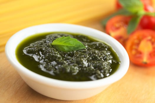 Fresh pesto made of basil, garlic and olive oil in a small bowl and garnished with a basil leaf with raw spaghetti, cherry tomatoes and basil leaves in the back (Selective Focus, Focus on the basil leaf in the bowl)