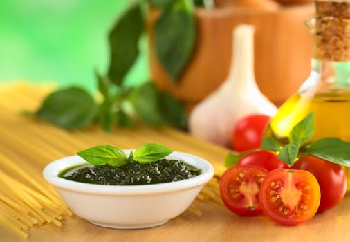 Fresh pesto made of basil and cherry tomatoes with olive oil, garlic, mortar, basil and raw spaghetti in the back (Selective Focus, Focus on the basil leaf on the pesto and the tomato in the front)