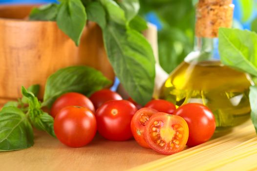 Cherry tomatoes on wooden board with other ingredients such as olive oil, spaghetti and basil with a wooden mortar in the back (Selective Focus, Focus on the first tomato half)