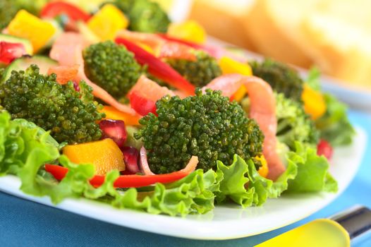 Fresh broccoli salad with lettuce, bell pepper, ham, mango and pomegranate with baguette slices in the back (Selective Focus, Focus on the broccoli floret in the front)