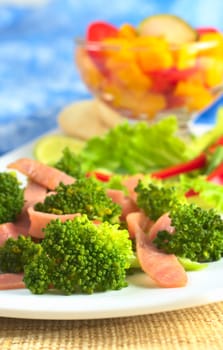 Fresh broccoli-ham salad with a bowl of mango and red bell pepper in the back (Selective Focus, Focus on the broccoli floret and the ham in the front)