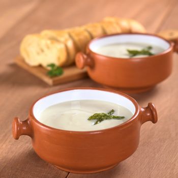 Cream of asparagus with green asparagus heads in rustic bowl with slices of baguette white bread in the back on wood (Selective Focus, Focus on the asparagus head in the first bowl)