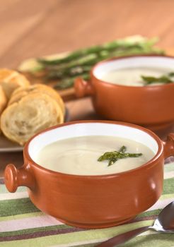 Cream of asparagus with green asparagus heads in rustic bowl with slices of baguette white bread and asparagus in the back on wood (Selective Focus, Focus on the asparagus head in the first bowl)