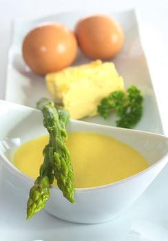 Asparagus lying on a bowl full of Hollandaise sauce with its ingredients, egg and butter in the back (Very Shallow Depth of Field, Focus on the head of the right asparagus)