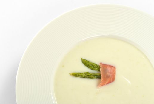 Cream of asparagus garnished with green asparagus heads and a slice of ham on top in a white soup plate (Selective Focus, Focus on the asparagus heads and the ham slice on the soup)