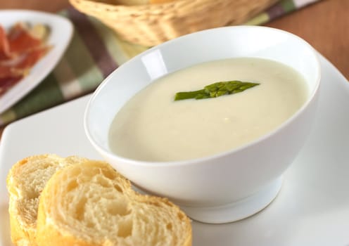 Cream of asparagus with a green asparagus head on top in a white bowl with baguette slices beside (Very Shallow Depth of Field, Focus on the asparagus head in the soup)