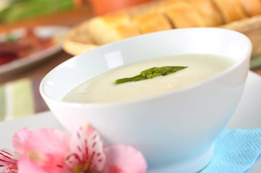 Cream of asparagus with a green asparagus head on top in a white bowl with a pink Inca Lily beside and baguette in the back (Very Shallow Depth of Field, Focus on the asparagus head in the soup)