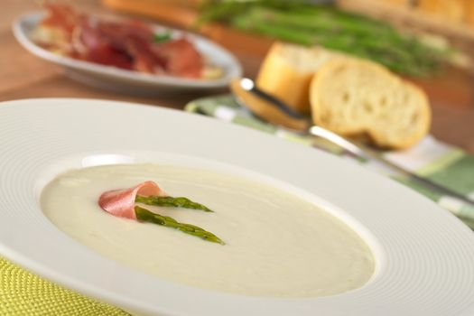 Cream of asparagus garnished with green asparagus heads and a slice of ham on top in a white soup plate (Selective Focus, Focus on the asparagus head in front and part of the ham slice on the soup)