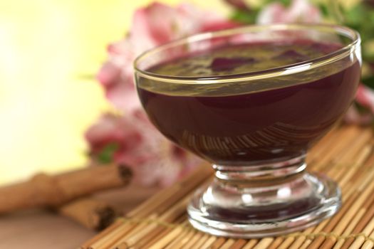 Popular Peruvian dessert called Mazamorra Morada (made out of purple corn) with cinnamon sticks and pink inca lily in the back (Selective Focus, Focus on the front of the bowl)