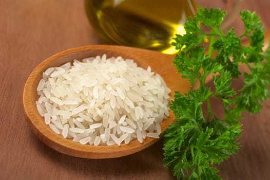 Raw rice grains on a wooden spoon with parsley on the side and oil behind (Selective Focus, Focus on the middle and the front of the grains)