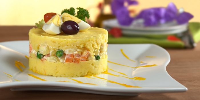 Traditional Peruvian dish called Causa made of mashed yellow and white potato mixed with aji (hot pepper) and lime juice and filled with vegetables (corn, peas, carrots) and mayonnaise and garnished with eggs and olives (Selective Focus, Focus on the front) 
