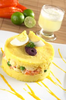 Traditional Peruvian dish called Causa made of mashed yellow and white potato mixed with aji (hot pepper) and lime juice and filled with vegetables (corn, peas, carrots) and mayonnaise and garnished with eggs and olives, with Pisco Sour in the back (Selective Focus, Focus on the front) 