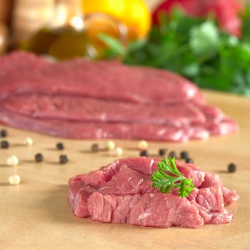 Fresh raw beef meat with parsley on top, with vegetables, herbs and kitchen utensils in the back (Selective Focus, Focus on the front of the meat and the parsley)