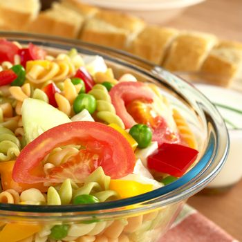 Pasta salad with fresh vegetables (tomato, pea, bell pepper, cucumber) and cheese with baguette and salad dressing in the back (Selective Focus, Focus on the tomato) 
