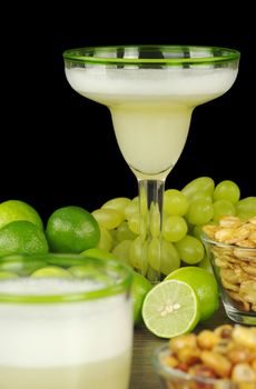 Pisco Sour, a Peruvian cocktail made of pisco, lime juice, sugar syrup and egg white. The cocktail is surrounded by grapes, limes, and Peruvian snacks, habas and canchas. (Selective Focus, Focus on the glass in the back)