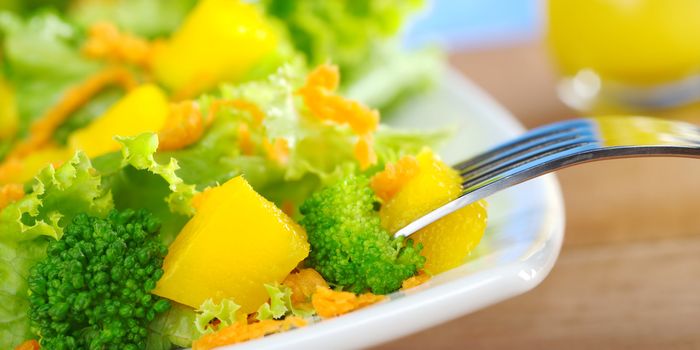 Fresh and light broccoli-mango-carrot salad on white plate with a piece of mango on fork and a glass of orange juice dressing in the background (Very Shallow Depth of Field, Focus on the mango and broccoli in the front and the front of the fork)