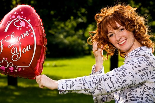 Red-haired lovely young woman holding red balloon in shape of heart outdoors 