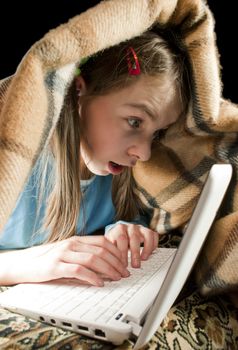 Teen girl with laptop surfing Internet and hiding under blanket