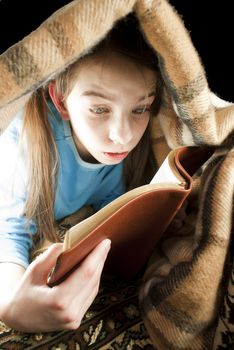 Teen girl reading book hiding under blanket