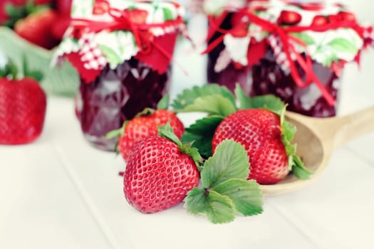 Fresh strawberries with jars of freshly preserved jam in the background. Selective focus on large strawberry beside of spoon.