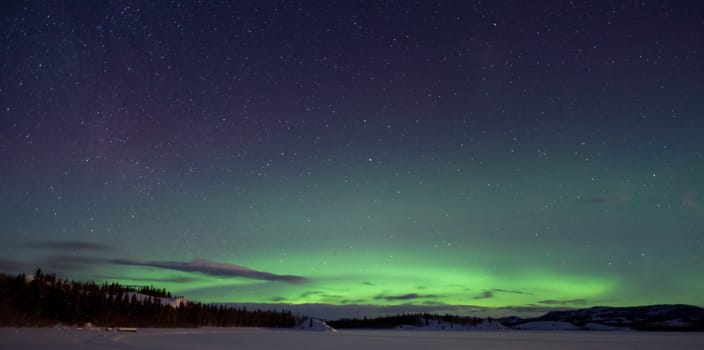 Green northern lights (aurora borealis) substorm, a few clouds and lots of stars on night sky above snowy hills.