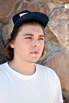 A young man smoking a cigarette wearing a white t-shirt and a trucker style hat.