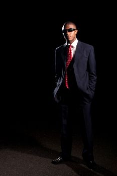  An African American man dressed in a dark colored suit and sunglasses standing in front of a dark black background.