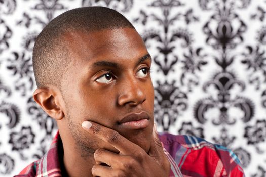 An African American man with his hand on his chin thinking deeply about something in front of a damask style background. Shallow depth of field.