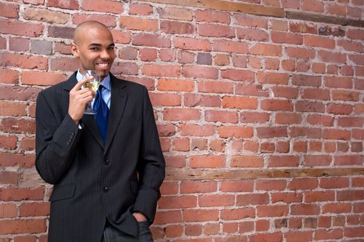 Young business man leaning against the brick wall, drinking wine and smiling.