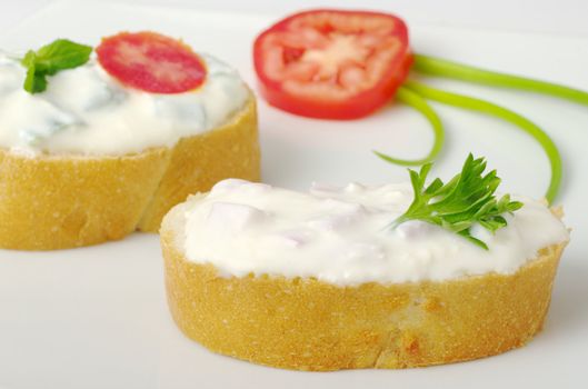 Baguette bread with cream cheese spread decorated with tomato, parsley and green onion on white plate (selective focus)